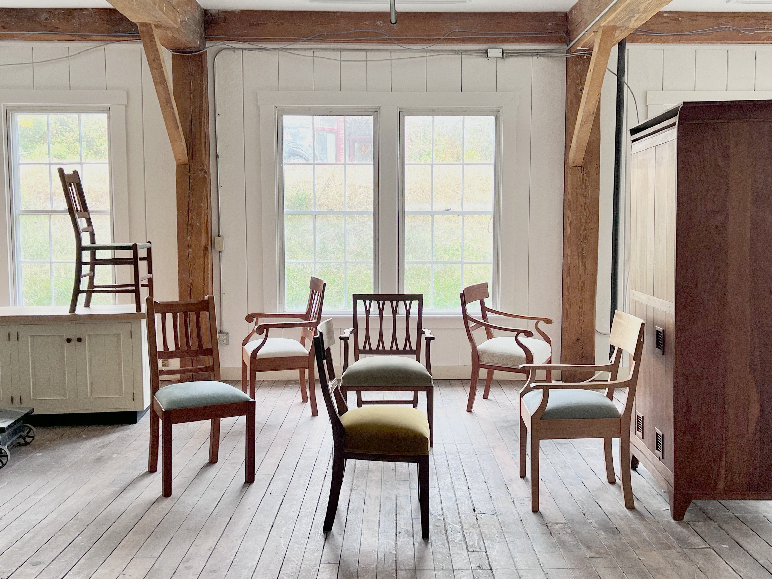 a group of wooden dining chairs in cherry, walnut, and maple in an empty space with large windows, beams, and wood floors.