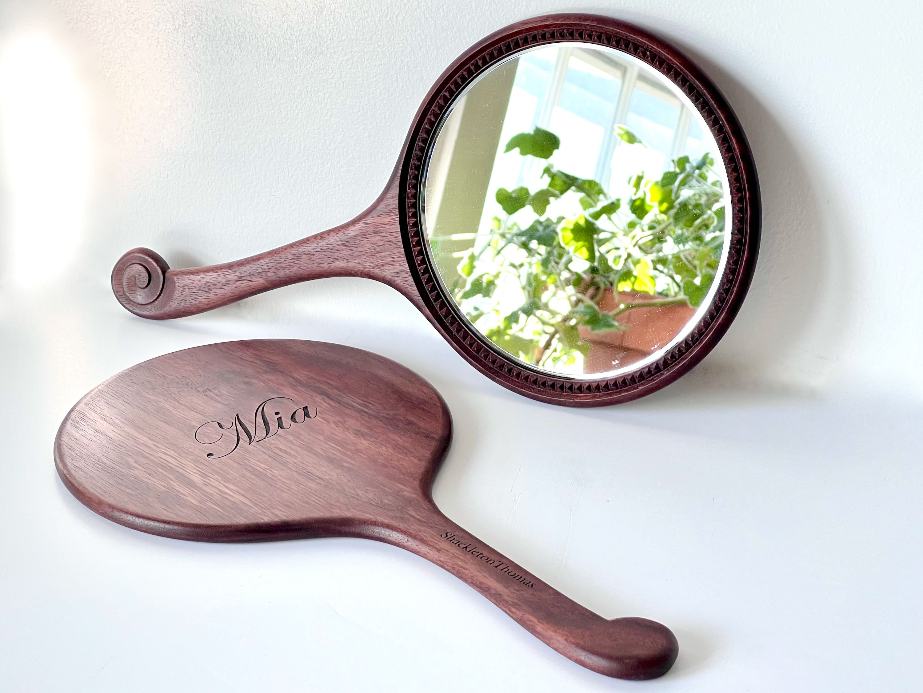 A hand-carved wooden mirror with curved molding and beveled glass, displaying a reflective surface with a plant in the background.