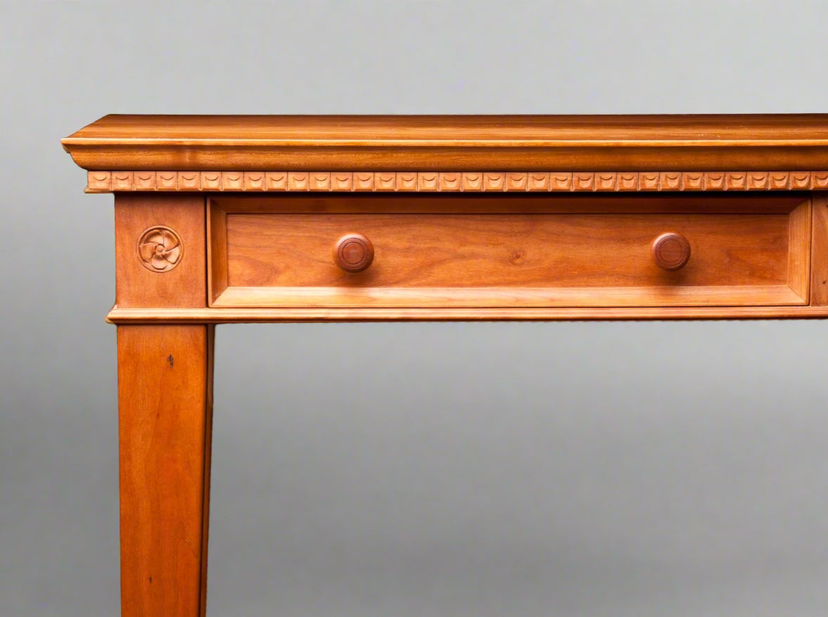 A classic wooden desk with a rectangular tabletop and carved details, including scalloped dentil molding and tapered legs.